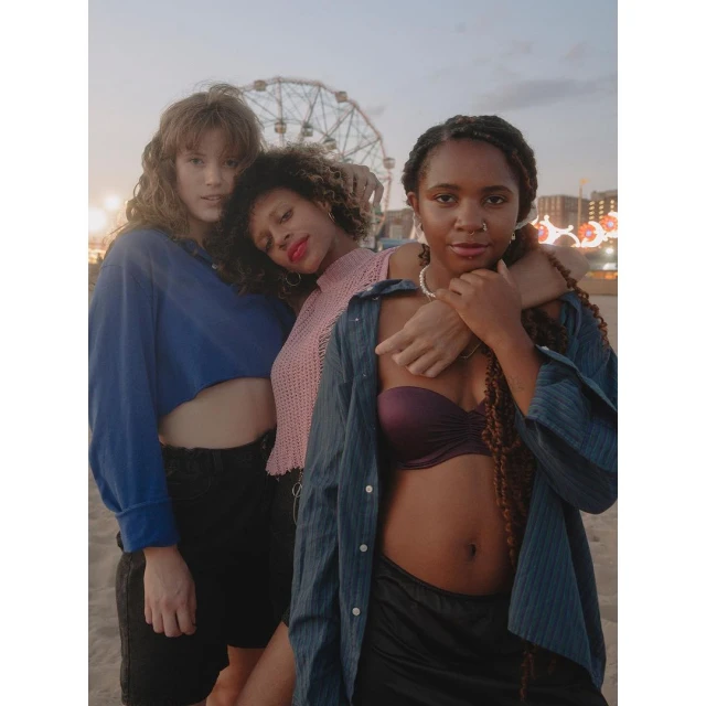Three young women pose on a beach at dusk, with a Ferris wheel visible in the background. They wear casual tops and shorts, with relaxed expressions.