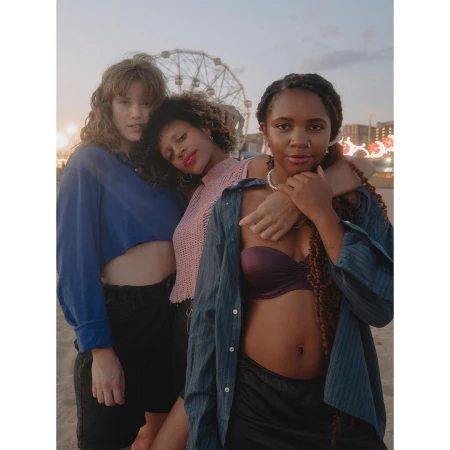 Three young women pose on a beach at dusk, with a Ferris wheel visible in the background. They wear casual tops and shorts, with relaxed expressions.