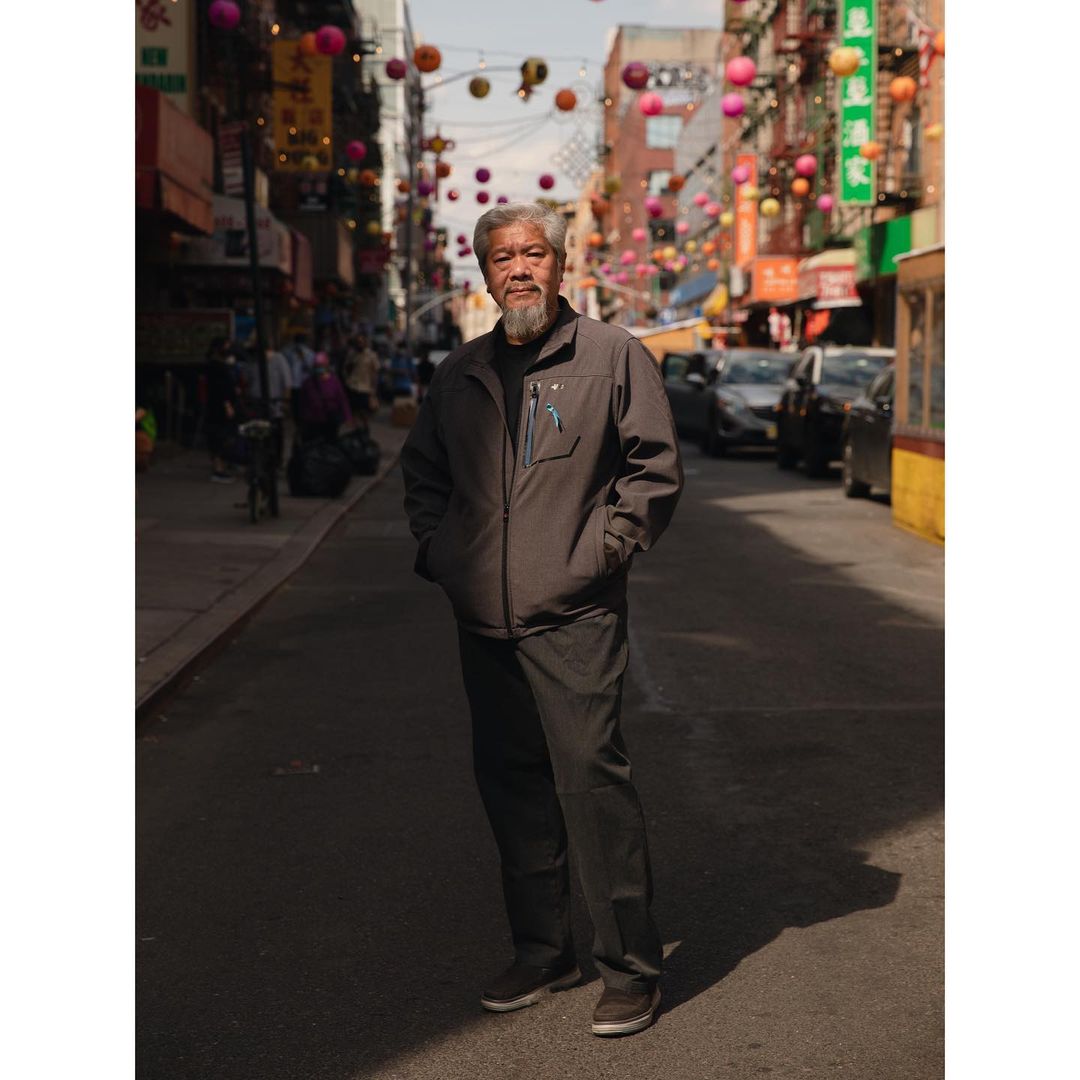 Elderly Asian man in a gray jacket stands on a city street surrounded by colorful hanging lanterns in an urban setting, capturing a street style moment.