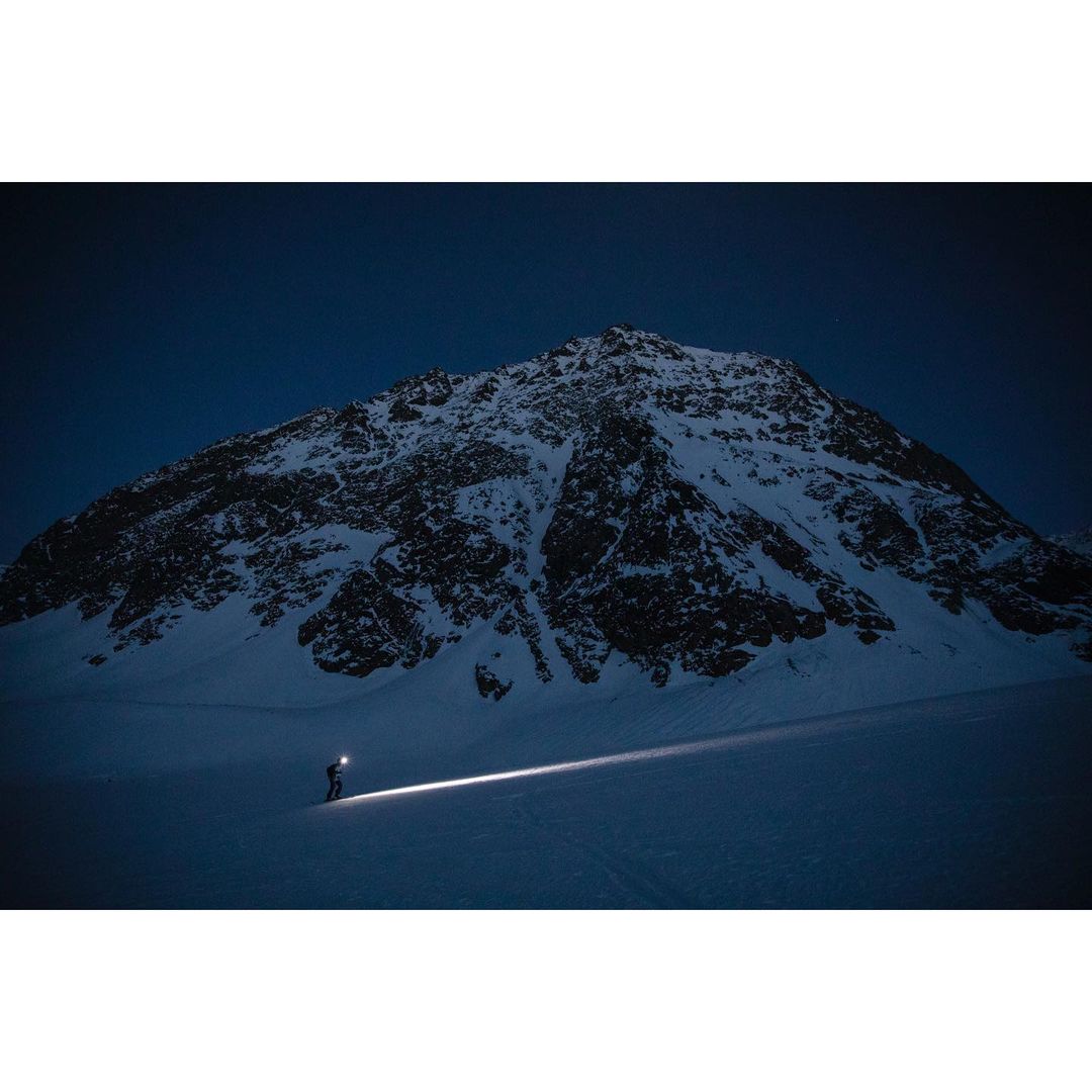 Person with headlamp ascends snowy mountain at night, creating dramatic light beam; landscape offers a sense of adventure and endurance.