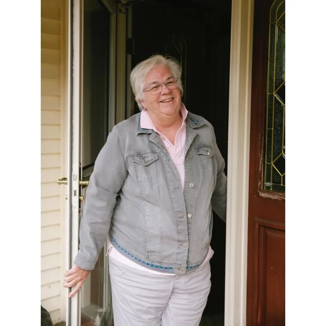 Mature woman stands in a doorway wearing a denim jacket, pink shirt, and striped pants, smiling towards the camera.