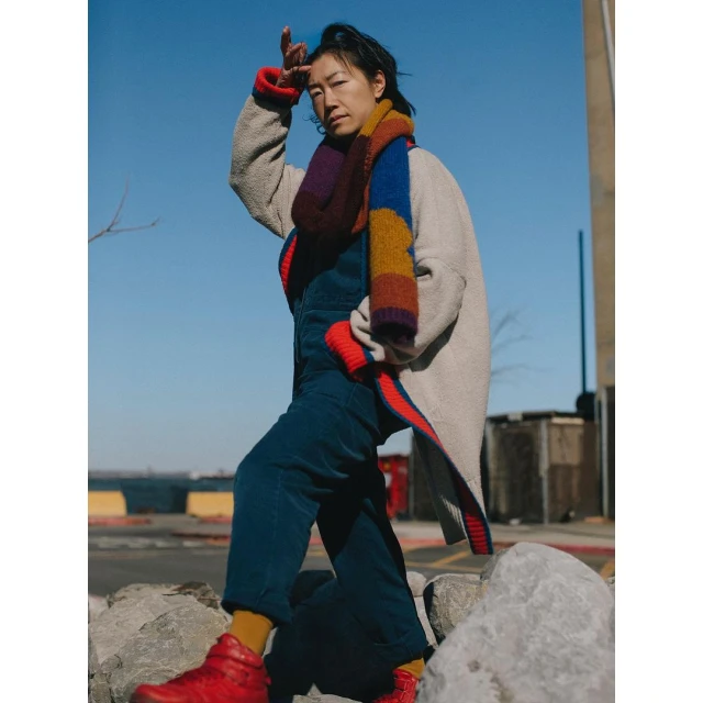 A woman in a beige coat, blue overalls, and red shoes stands on rocks against a clear blue sky in an outdoor urban setting.