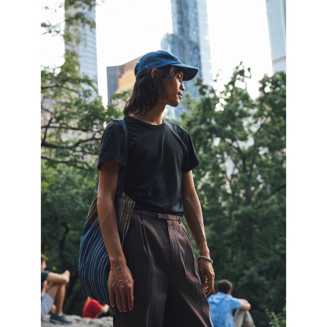 A young adult in a black t-shirt and blue baseball cap, carries a tote bag in an outdoor urban setting with skyscrapers.