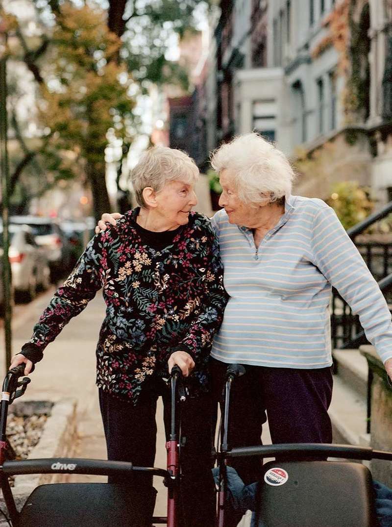 Two elderly women with walkers enjoy a walk on a tree-lined city street, wearing casual clothes and smiling at each other in daylight.