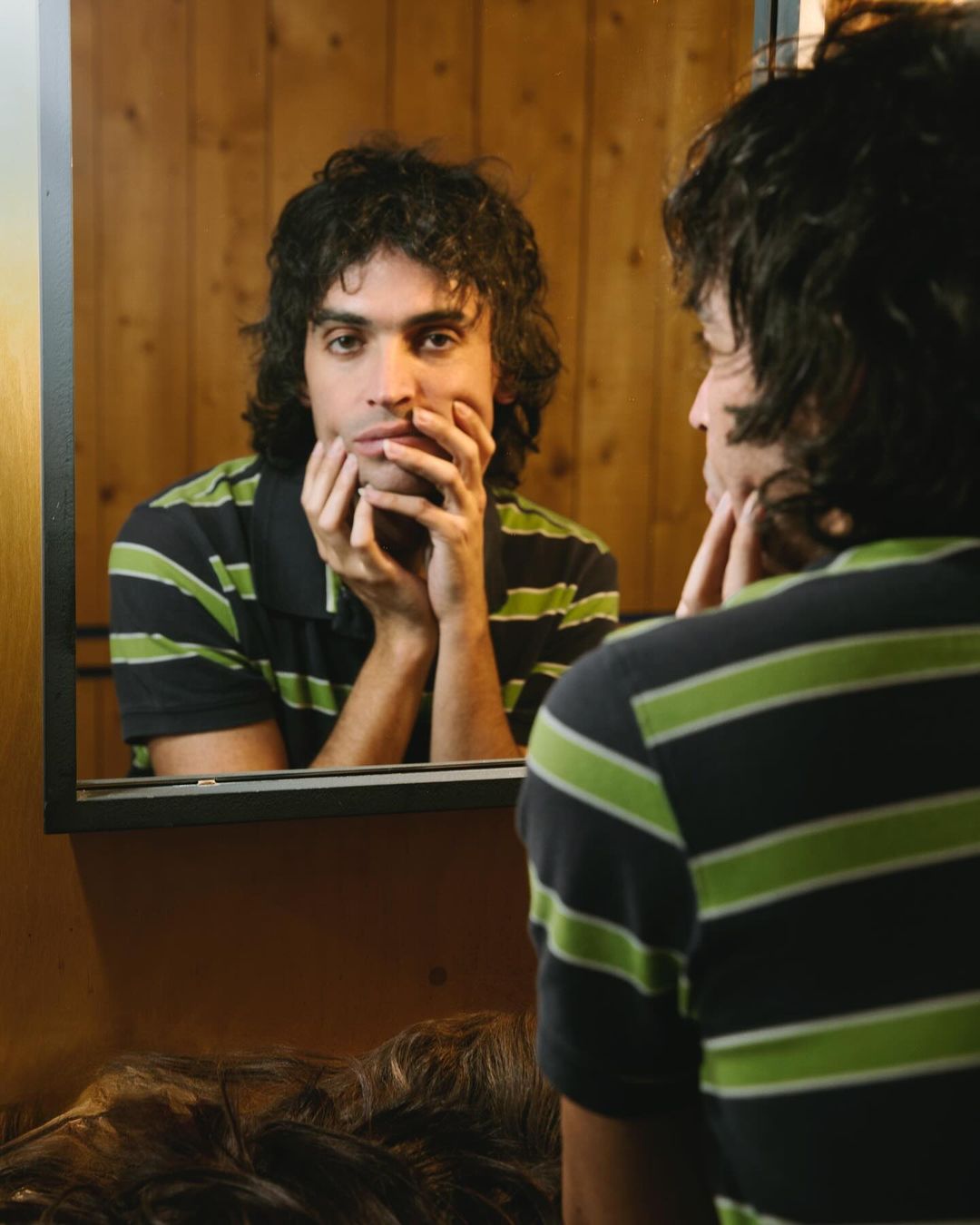 Man gazes into a mirror, his face reflected with hands near his cheeks, wearing a striped polo shirt indoors with wood paneling.