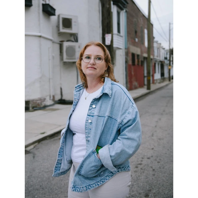 Woman with auburn hair wears eyeglasses and a denim jacket, standing on a city street lined with buildings and utility fixtures.