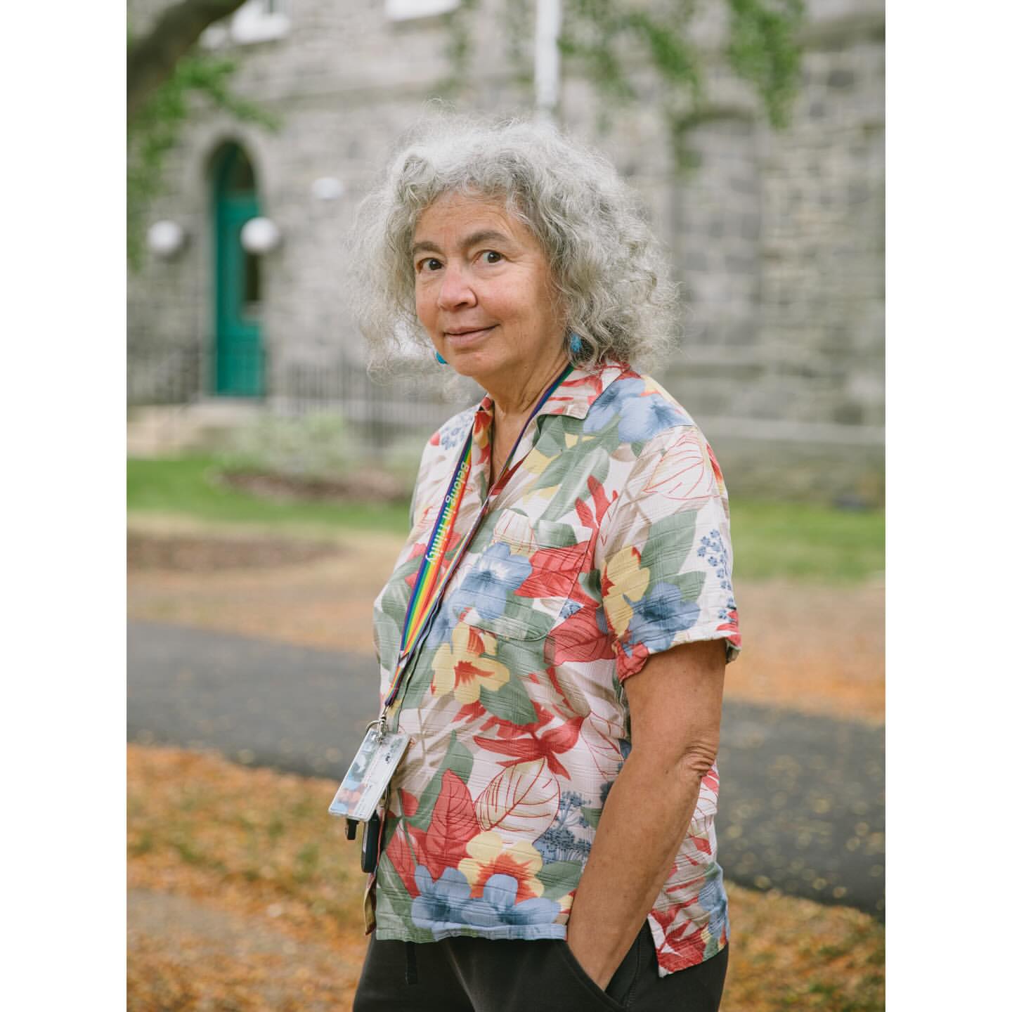 Portrait of an elderly woman with curly gray hair, wearing a floral shirt and lanyard in an outdoor setting with a stone building.