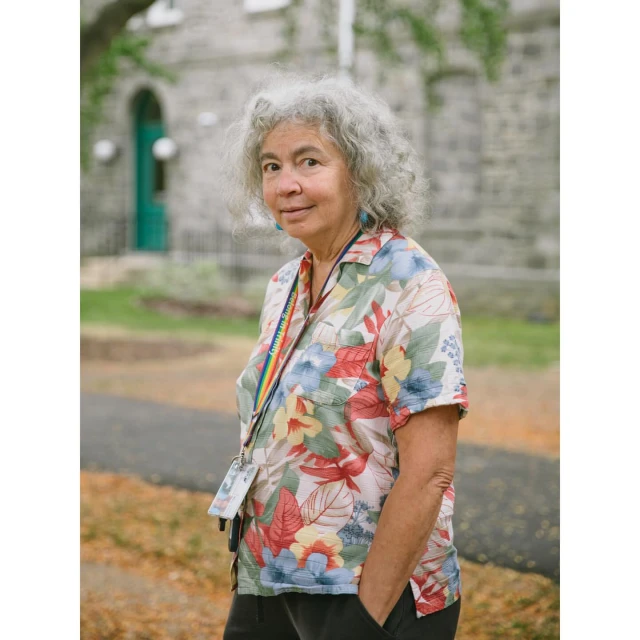 Portrait of an elderly woman with curly gray hair, wearing a floral shirt and lanyard in an outdoor setting with a stone building.