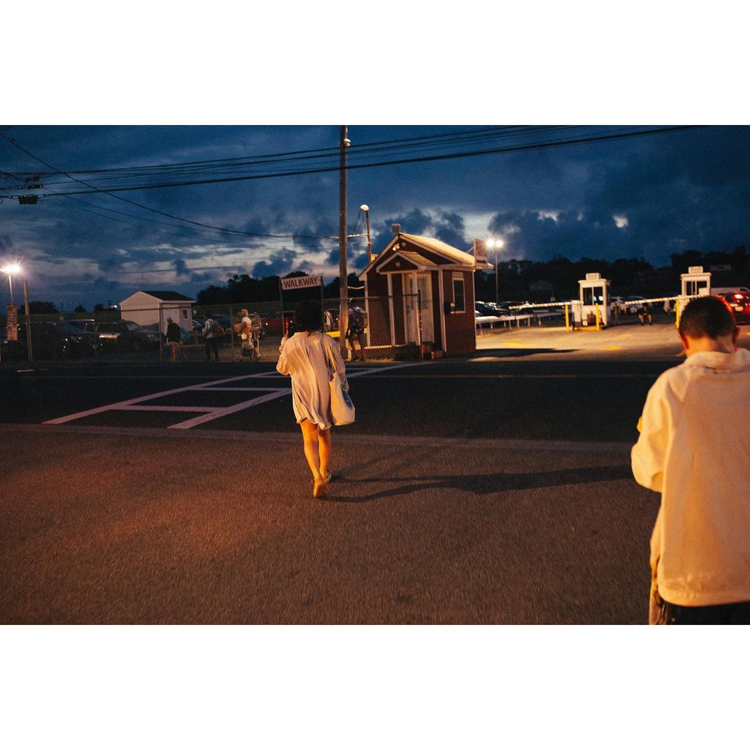 Woman in a white shirt walks on a crosswalk towards a red building at night with dark blue sky and some light poles visible.