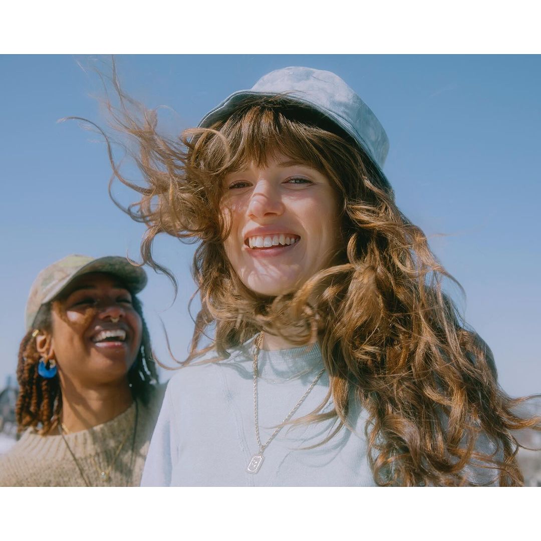 Two women pose outdoors on a sunny day. One woman is smiling and looks directly at the camera. The second woman is smiling.