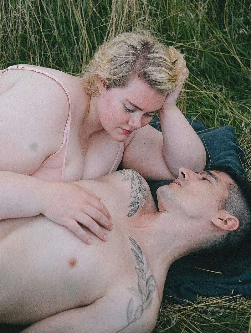Couple lying in grassy field, relaxed pose. Woman with blonde hair gazes at man with dark hair and tattoos, nature background.