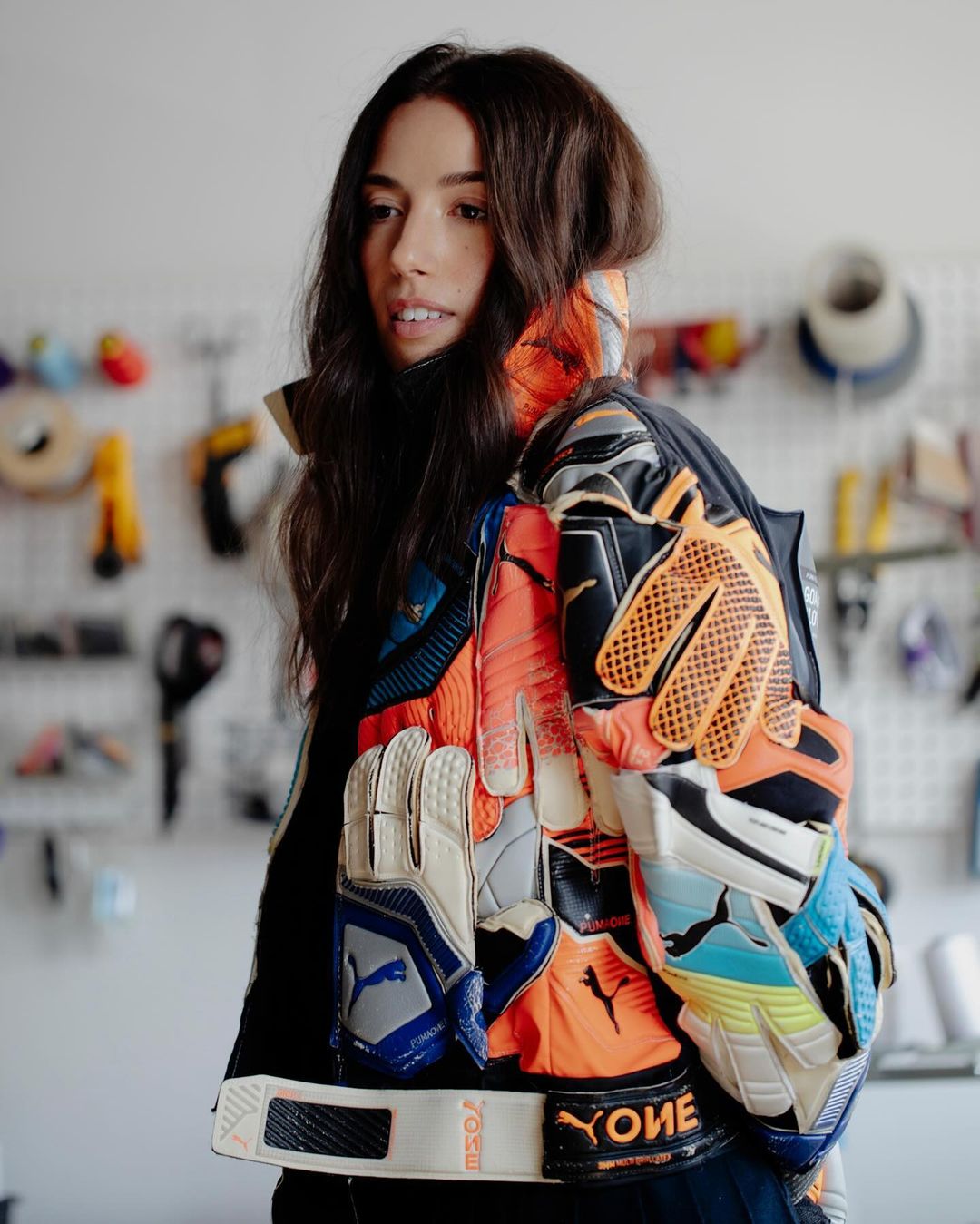 Brunette woman wearing a jacket made of colorful goalie gloves in an indoor setting with various tools in the background.