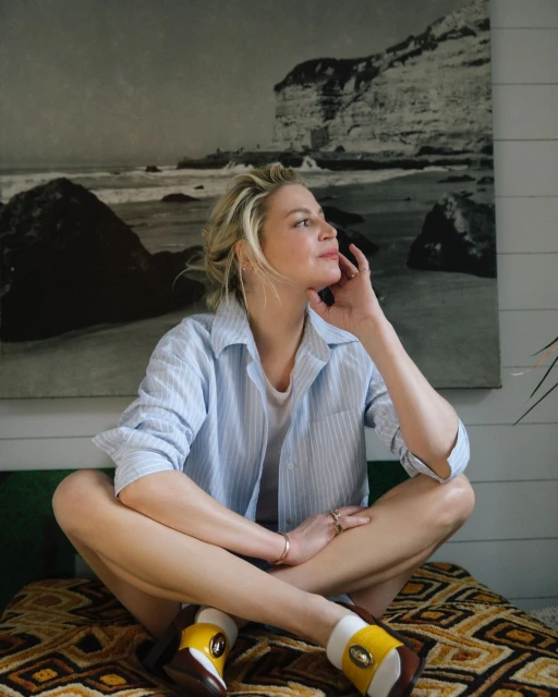 Woman sitting cross-legged indoors with a black-and-white beach scene art print and wearing a blue striped shirt and white shoes with yellow accents.