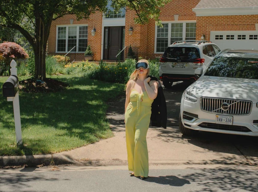 A blonde woman in a yellow jumpsuit and ski goggles stands on a street near two cars and a brick house in a suburban area.