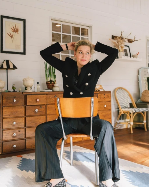 Woman in blazer and wide-leg pants poses seated on a chair in a light-filled interior with hardwood dresser and patterned rug.