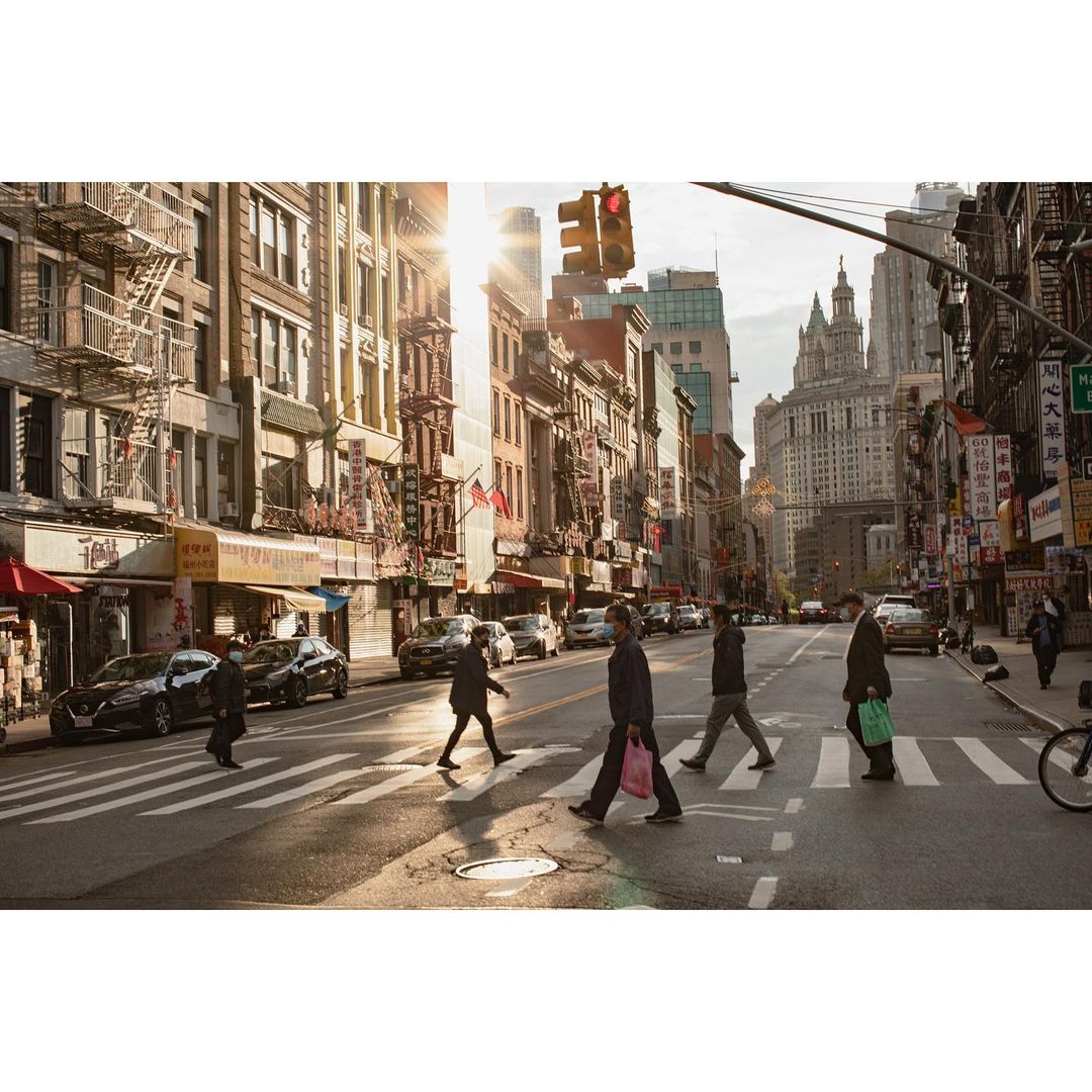 Street view of pedestrians crossing a zebra crossing in New York City, with buildings and cars visible under bright sunlight.