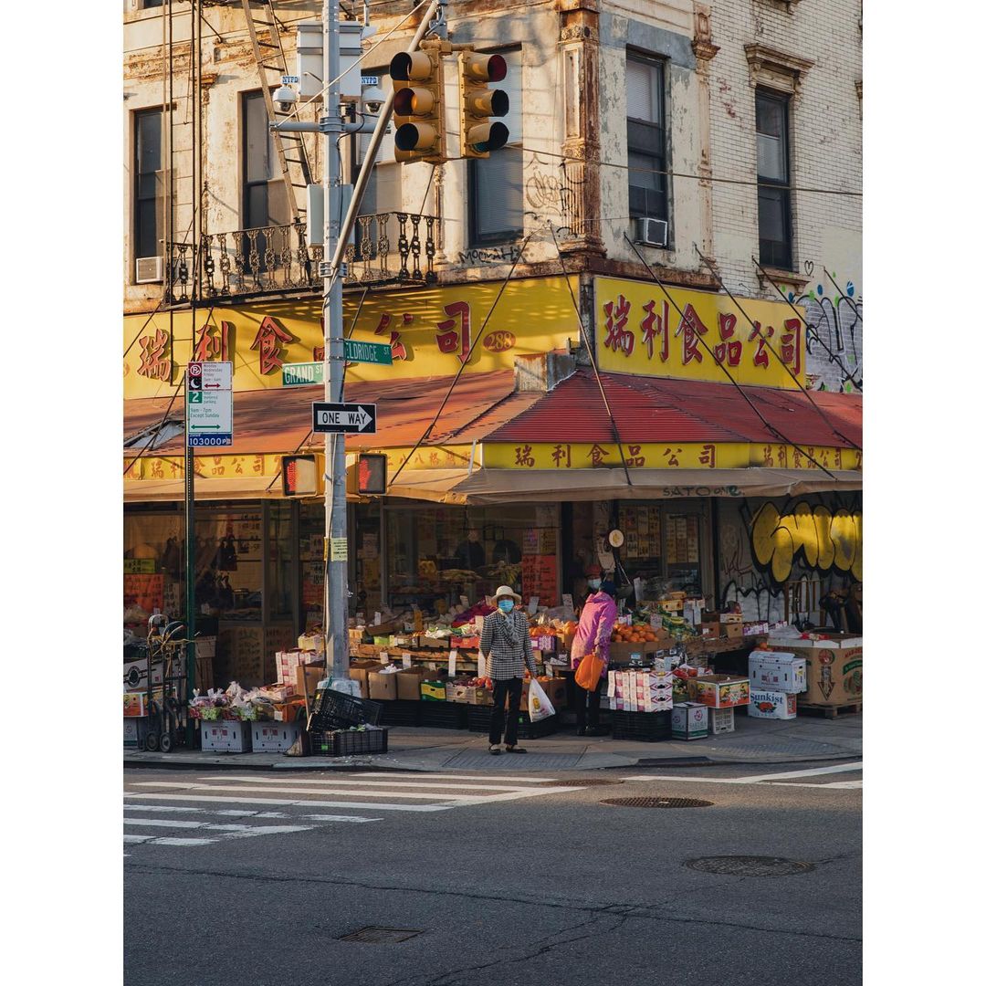 Street corner view of a yellow-awninged market with two women in masks, surrounded by fruit and vegetable displays on a sunny day.