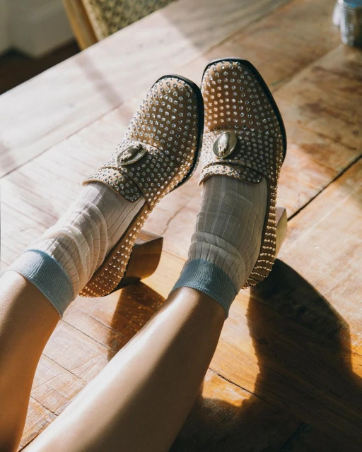 Close-up of studded loafer-style heels on a wooden floor, worn with ribbed socks, legs angled toward sunlight, creating shadows.