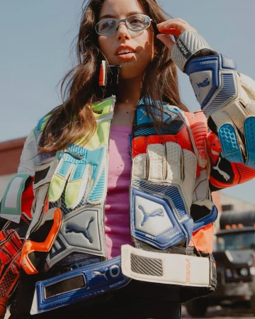 Portrait of a woman wearing a jacket made of repurposed goalkeeper gloves, pink shirt, and glasses, standing in an urban environment.