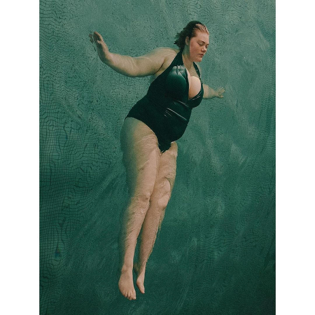 Woman in a black swimsuit swimming in water. The image captures the model underwater with sunlight filtering through the surface.