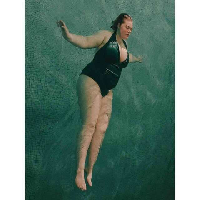 Woman in a black swimsuit swimming in water. The image captures the model underwater with sunlight filtering through the surface.
