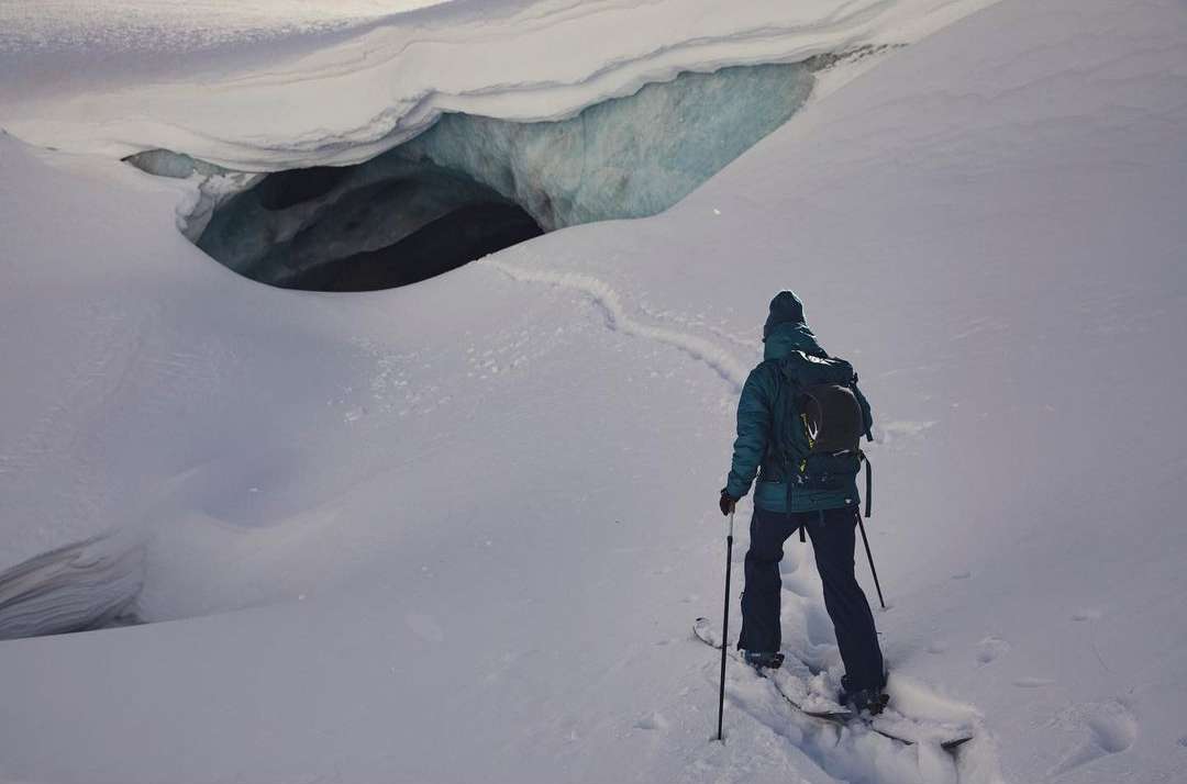 Person with backpack on skis approaches a snow cave entrance in a winter landscape, offering an adventurous outdoor scene.