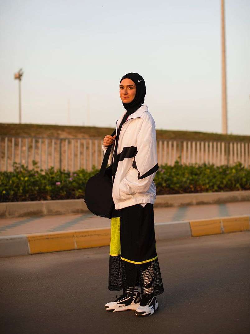 Female athlete in Nike activewear stands on an outdoor track. She wears a black hijab, jacket, long skirt, sneakers, and shoulder bag.