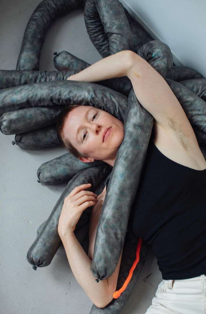 Artistic portrait of a woman posing with soft, gray sausage-like objects, indoor setting against a simple wall, neutral color palette.