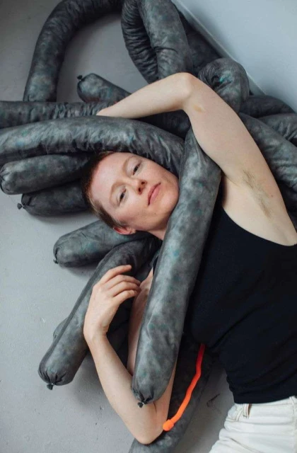 Artistic portrait of a woman posing with soft, gray sausage-like objects, indoor setting against a simple wall, neutral color palette.