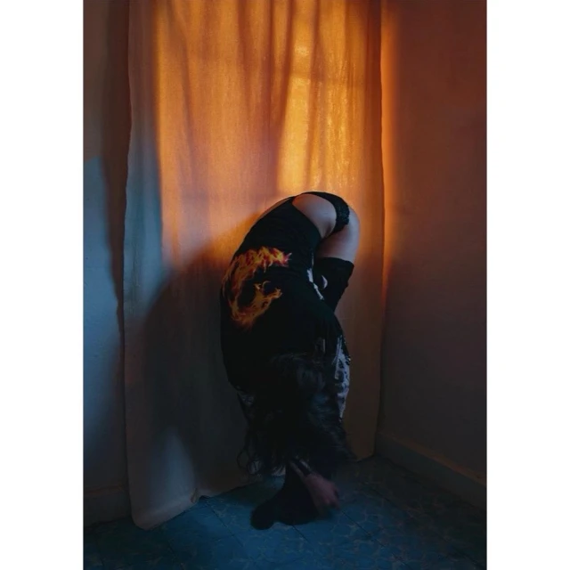 A woman bends over in a room with a vintage aesthetic, wearing dark clothing, with light from a window illuminating the scene.