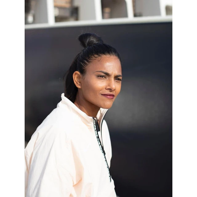 Woman with bun hairstyle in light pink jacket poses outdoors against a dark background in portrait orientation.