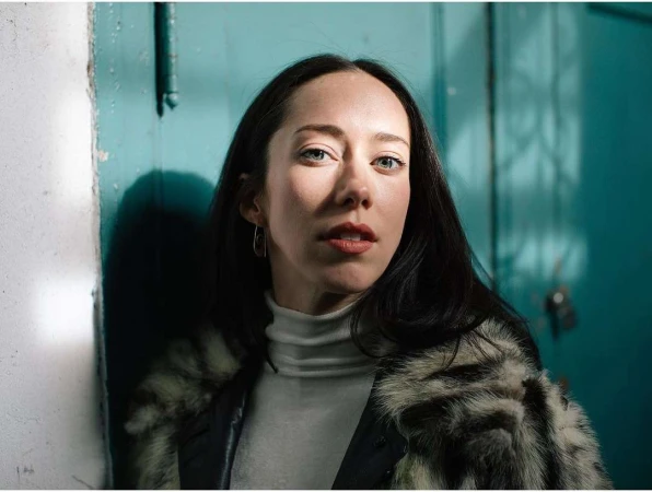 Portrait of a woman with dark hair in a white turtleneck and patterned jacket against a teal door