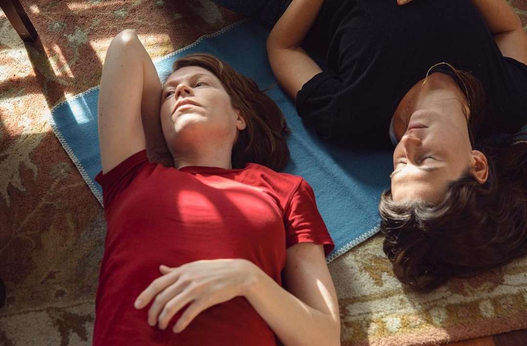 Two women lie on a blue mat, one in a red shirt looking up, the other in a black shirt with eyes closed, indoors on a sunny day.