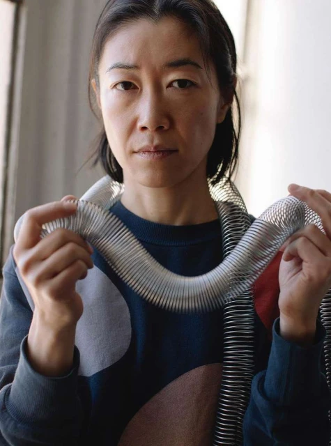 Portrait of an Asian woman indoors holding a large metal slinky toy and looking toward the camera with a neutral expression on her face.