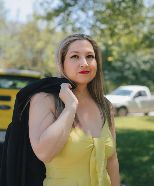 Outdoor portrait of a smiling woman in a yellow dress with a black jacket over her shoulder, set against a natural green background.