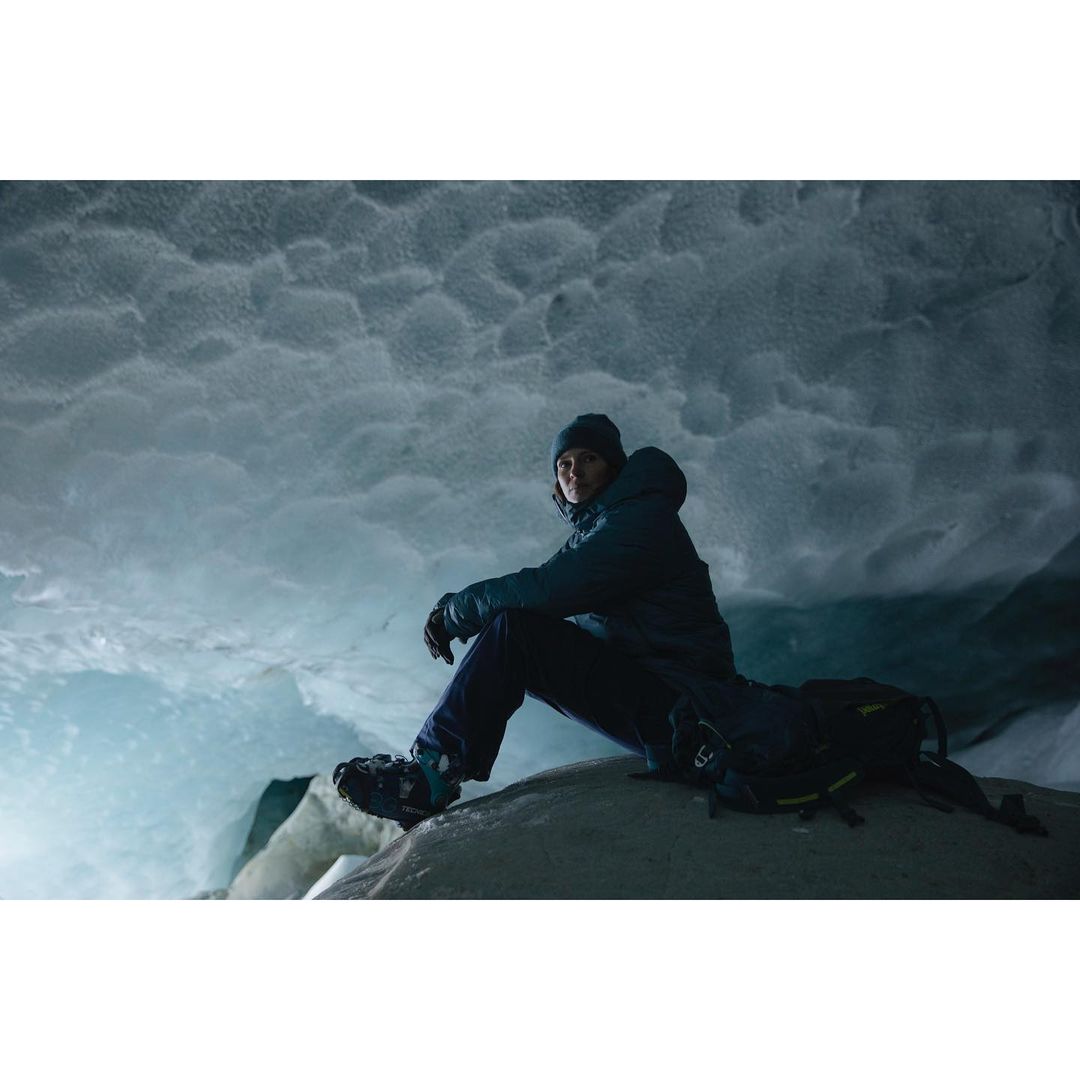 Adult in a jacket and beanie sits on a rock with a backpack inside of a glacier cave featuring blue ice and textured formations.