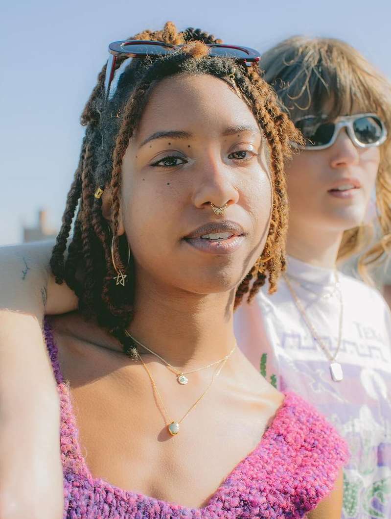 Two women pose outdoors in a medium shot, one in a pink top and the other in white, both wearing sunglasses in the sunlight.