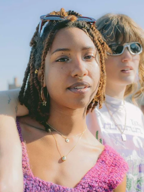 Two women pose outdoors in a medium shot, one in a pink top and the other in white, both wearing sunglasses in the sunlight.