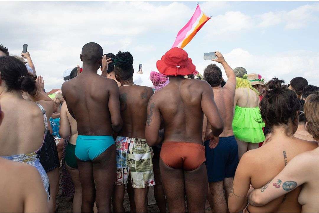 Crowd of people gathered at the beach during daytime, with some individuals wearing swimwear and holding phones, and a flag in the background.