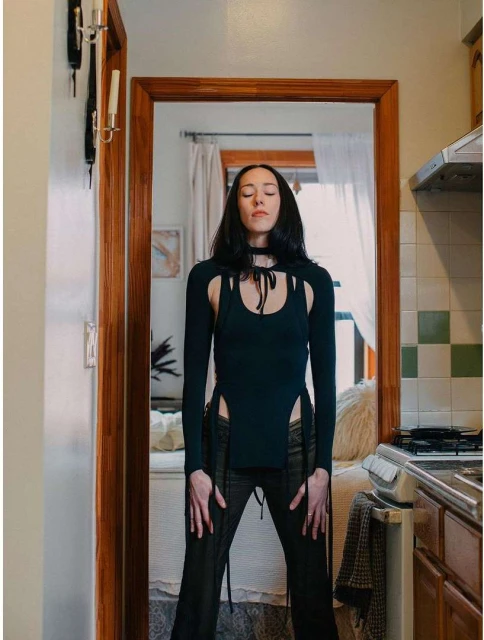 Woman stands in black cut-out top and pants, eyes closed, with a bed and kitchen visible in the background in an indoor setting.