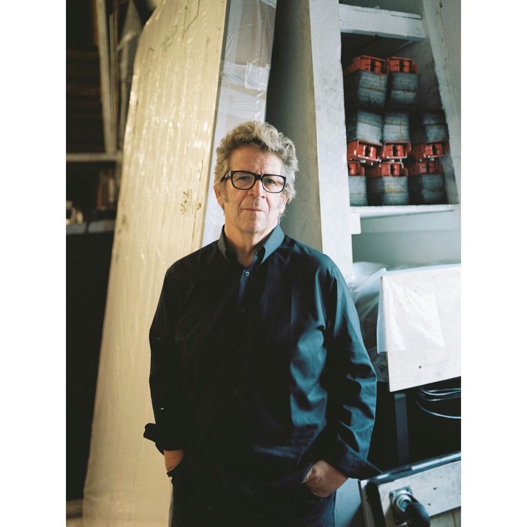 Adult male with eyeglasses wearing a dark button-down shirt posing indoors near large wrapped architectural elements and stacked red objects.
