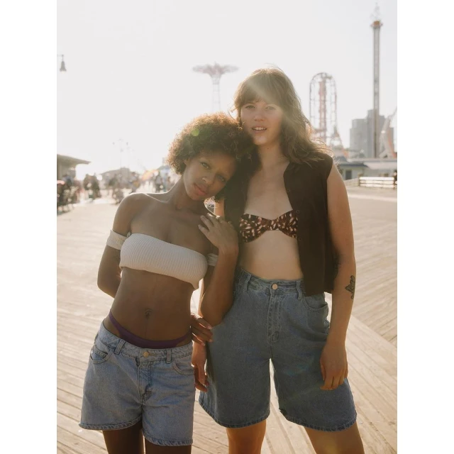 Two women in casual summer outfits pose on a boardwalk in bright sunlight with amusement park rides visible in the background.