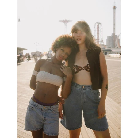Two women in casual summer outfits pose on a boardwalk in bright sunlight with amusement park rides visible in the background.