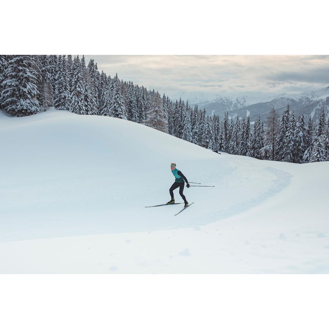 Person cross-country skiing on snow-covered field lined with trees on a cloudy day in a mountain landscape. Sport and winter scenes.