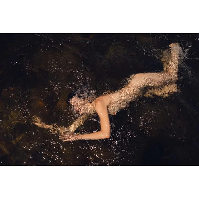 Overhead underwater photograph of a woman swimming in dark water over submerged rocks by Katharina Poblotzki