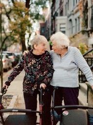 Two elderly women smiling at each other while walking down a city street using rolling walkers