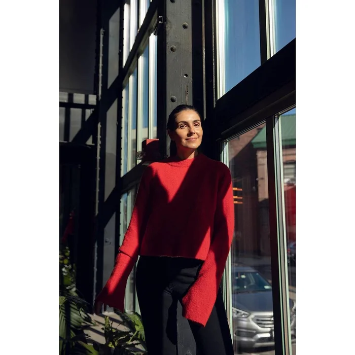 Woman wearing a red crewneck sweater standing in front of large industrial windows by Katharina Poblotzki