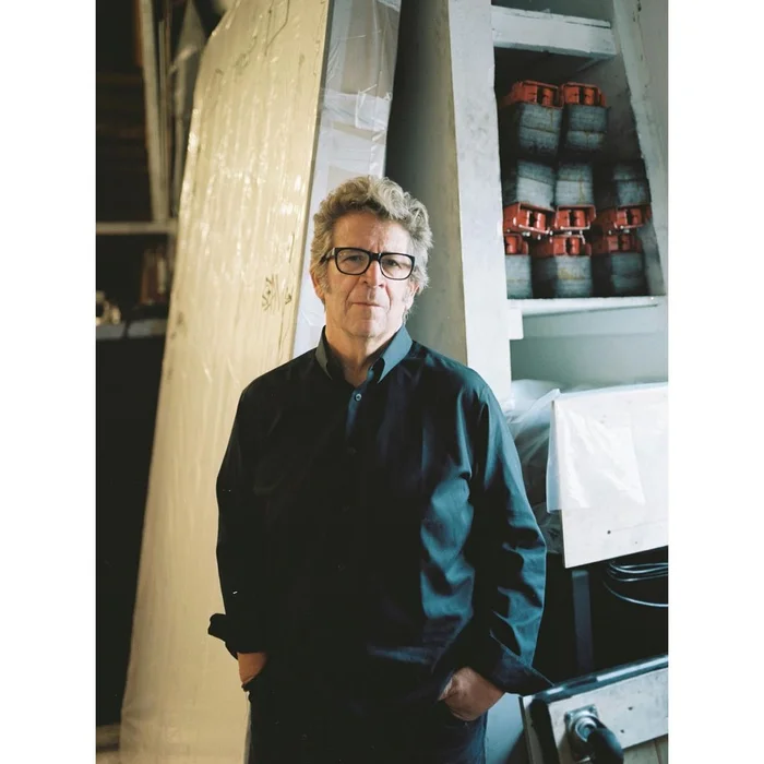 Portrait of an older man wearing glasses and a dark shirt standing in a workshop or studio setting by photographer Katharina Poblotzki