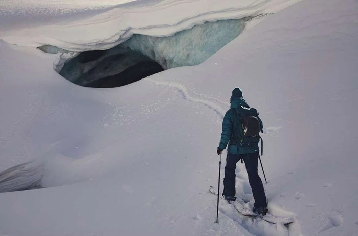 Skier descending past a dark glacial ice cave in a vast snowfield under cool natural light