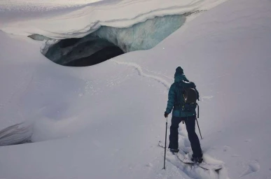 Skier descending past a dark glacial ice cave in a vast snowfield under cool natural light
